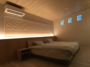 寝室、説明キャプション：こちらもお気に入りの寝室。間接照明によって、天井の木目がグラデーションに。リクシルの壁材「エコカラット」の凹凸も際立つ